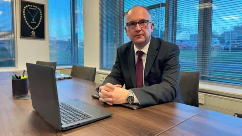 A man is sat down inside an office wearing a black suit, white shirt and red tie. In front of him is a laptop. 