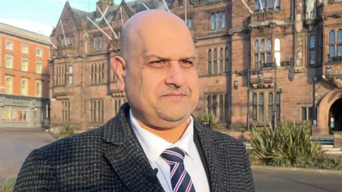 Abdul Khan stands outside Coventry Council House. He wears a grey chequered coat and a blue tie with red stripes.