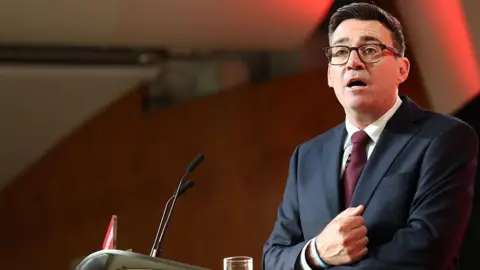 Andy Burnham wearing a black jacket with a black t-shirt talking to an audience with a black background behind him