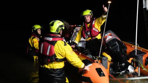 RNLI volunteers on an inshore lifeboat which is in the water making its way to an incident. One of the men has put his thumb in the air, directing two other volunteers in the water.