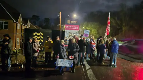 Sharna Maria A group of 20 people with banners protesting about an army camp being used to house asylum seekers stand on a pavement in the dark.