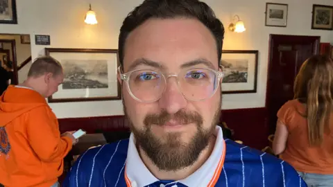 Ollie Kay wearing a blue grey and orange striped Luton Town football shirt. He is standing in a pub.