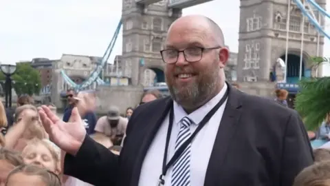 Joshua Falcon/BBC A man wearing a black suit blazer over a white shirt with a striped tie stood in the middle of a group of schoolchildren who are wearing white and red uniforms. In the background is Tower Bridge.