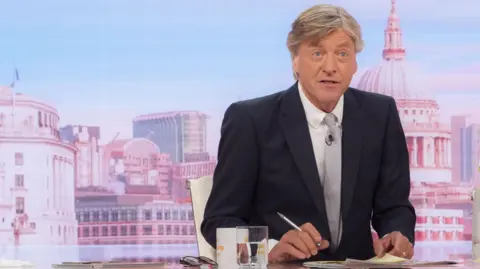 Shutterstock Richard Madeley sitting behind a desk presenting Good Morning Britain. He is wearing a dark suit jacket, white shite and grey tie. Behind him the dome of St Paul's Cathedral can be seen. He is holding a silver pen in his left hand. A mug, a glass of water and his spectacles are sitting on the desk in front of him.