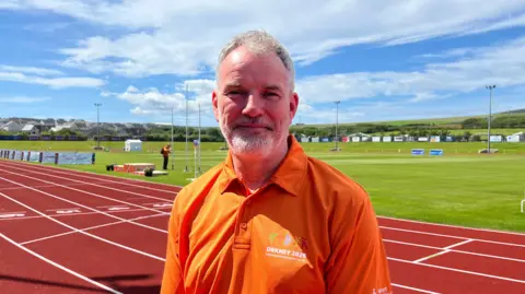 Gordon Deans is standing at The Pickaquoy sports centre in Orkney. He is stood on the athletics track with a field behind him. He is wearing an orange polo shirt with Orkney 2025 branding on it and smiling at the camera.