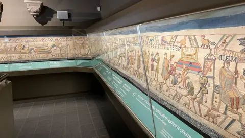The Bayeux Tapestry depicting the Norman conquest in 1066 is pictured behind a glass case along a grey wall.