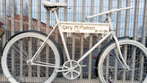 A bike painted white has been erected on railings with a sign saying Gary McMahon r.i.p 26-11-24