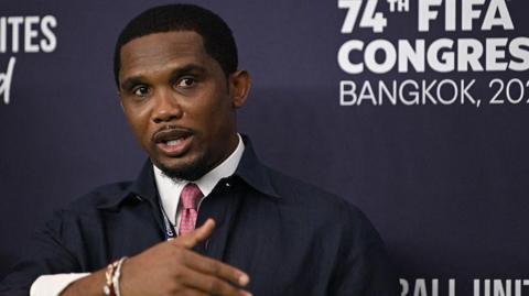 Samuel Eto'o gestures with his right arm while wearing a shirt and tie and standing in front of a media board that says 74th Fifa Congress Bangkok