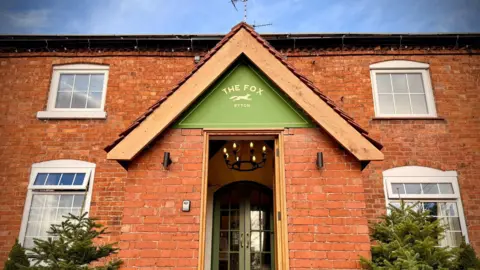 Steve Lee Exterior photo of the Fox Inn. It's a two storey, red brick building with four white framed windows visible. The front porch has a green sign over the door that reads 'The Fox, Ryton' with an illustration of a running fox. Either side of the porch are two Christmas trees. 