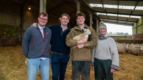 Ben, Hamish and Angus Weir are standing in a farm yard, with their mum Kathy. Angus is holding a lamb and there are many sheep in the background.