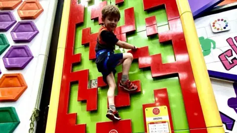 Liz Hardy Ben half way up a multi-coloured climbing wall. He has on shorts and a T-short and it looking at the camera.