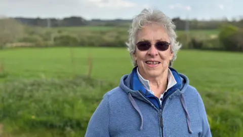 Kate has curly grey hair and round sunglasses. She wears a blue hooded top and stands in front of the fields where the solar farm will go. 