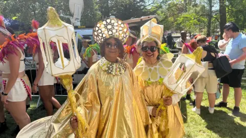 Lily-May Symonds/BBC Two participants wearing matching gold outfits pose for the camera. They wear long, gold robes complete with headdresses and matching staffs. Crowds surround them in a park during a sunny day.