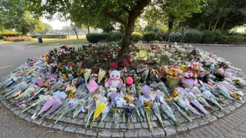Stuart Woodward/BBC Many flowers and soft toys have been left in a circle surrounding a small tree in the park. More trees and bushes are in the distance.