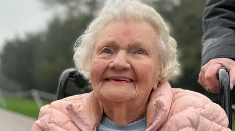 Sheila Levene, who has white hair, is wearing a pink coat and is sat in a wheelchair.