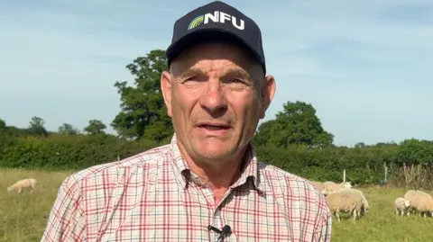 A farmer wearing a checked shirt and a cap with NFU written on the front. He is stood in a field with sheep and hedgerow behind him