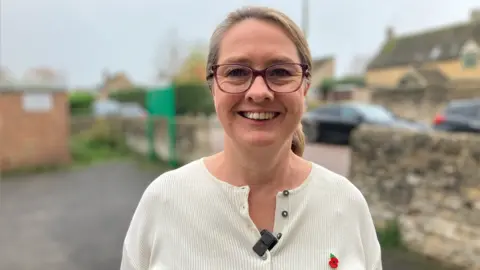 Alex Dunlop/BBC Helen Harrison, smiling and looking at the camera, with a brick wall and community centre behind her, slightly blurred. She has long fair hair tied back, is wearing glasses and a cream top, with a microphone on it and a poppy to the right. 