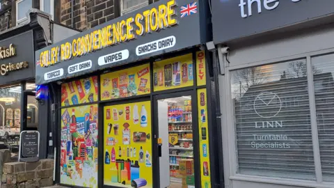 Otley Road Convenience Store. The shop has yellow posters in the windows with pictures of food and drink items.