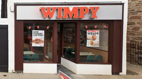 The Wimpy is in Dingwall High Street. The front is on the white-painted gable end of a peak roof building. There is a Wimpy sign above the door in red letters.