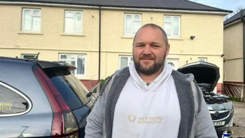 A middle-aged man wearing a white hoodie with a grey zip jacket on top. He has a serious expression on his face. He is stood in front of two silver parked cars, one with its bonnet up. Behind them are two attached houses. 