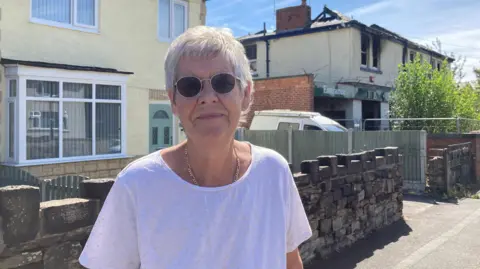 BBC/Naj Modak Susan Sheedy wearing round sunglasses and a white short-sleeved top standing on a residential street beside a low stone wall. In the background are houses, a green fence, a white van, and a fire-damaged building with broken windows and roof damage.