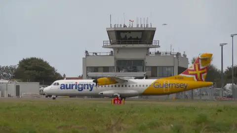 BBC White plane with a yellow tail with the words Aurigny and Guernsey on the side with a Guernsey flag on the tail wing. Behind it is the air traffic control tower. 