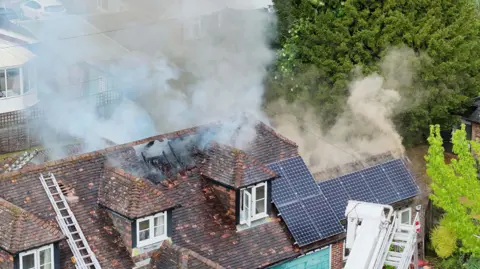 Eddie Mitchell A tile roof of a house, with solar panels. There is a hole in the top of the sloped roof, which has smoke billowing out.