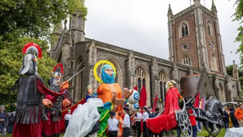 Toby Shepheard St Albans cathedral with a procession of people walking past, some wearing huge puppet costumes -Roman centurions, St Alban himself and a chariot. A group of children in scout hats and white t shirts stand around the saint puppet.