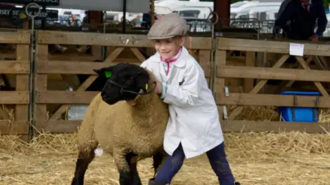 A girl holds and stands next to a sheep within a pen at the Suffolk Show that is filled with straw. She wears a white coat, flat cap and blue jeans with boots. The sheep has white wool and a black face and legs.