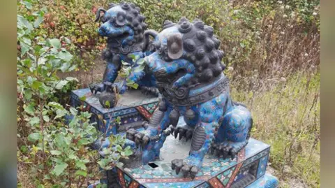 The two statues, pictures at the site. They are quite large, and are both of highly stylised lions covered with ornate carved and painted patterns. They are both sat on matching platforms, and have been found on an area of cracked concrete amongst high-grown weeds. 