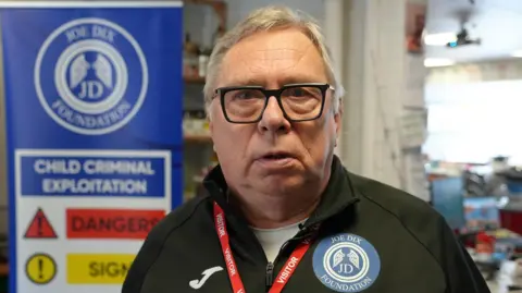 Qays Najm/BBC Alan Bliss, a man standing in front of a sign which has the Joe Dix Foundation branding on it. He is looking directly at the camera, with a pair of black framed glasses on and a black zip-up jacket.