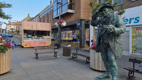 The bronze statue of Jimmy Stength is stood in the middle of a high street. A donut stall is stood near the benches to the left of the statue.