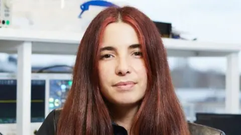 Carmen Palacios-Berraquero looking directly down the camera; only her head and shoulders are in frame. She has straight, dark red hair that falls below her shoulders. She is sitting inside a laboratory environment with some shelves and equipment behind her.