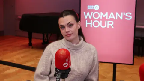 Charli sits in front of a microphone. She wears a beige jumper, her dark hair is pulled back from her face