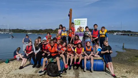 Romsey Young Carers A group of children sat on makeshift boat equipment next to a lake during a Romsey Young Carers activity day