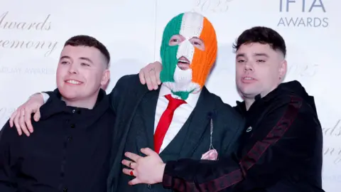 Reuters Three men are standing smiling for a photo. Left, is wearing a black coat and holding a glass. He is smiling at the camera and has short brown hair. The one in the middle is wearing a grey tartan suit and a red tie. He is also wearing a balaclava which is green white and orange striped. On the right, a man has his arms around the one in the middle. He is wearing a dark coloured jacket and has short black hair.