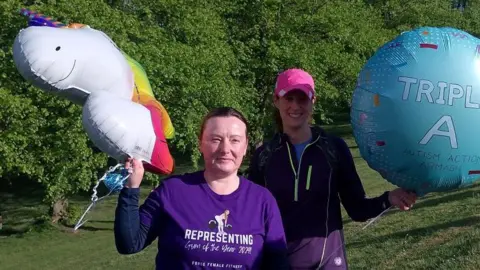 Jill Chadburn has dark hair pulled back in a ponytail. She is wearing a purple top with a navy long sleeved top under it. She is holding a white unicorn with a multicoloured tail. Beside her is a woman with a pink cap, blue top, black jacket and purple shorts holding a blue balloon.