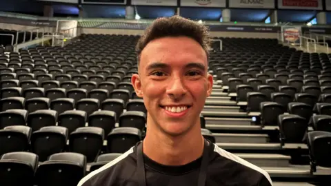 Youcef is smiling at the camera in a black and while tshirt. He is standing in front of the seating bank in the SSE Arena with his Riverdance lanyard around his neck.
