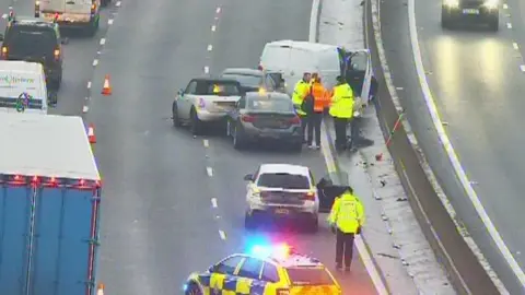 National Highways Traffic passes by a a multi-vehicle crash. People in hi-visibility jackets stand near the crash site