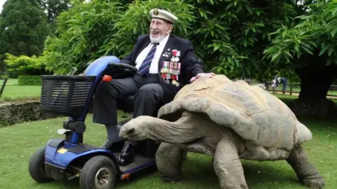 George Winter in full military regalia and riding an electric mobility scooter with his hand on a giant tortoise