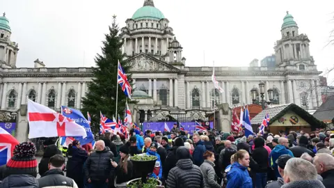Pacemaker A crowd gathered below Belfast City Hall, a large ornate building with featuring a prominent green copper dome at its center and towers at each of its four corners. A Christmas is tree and stalls are in front of it. The crowd are wearing winter attire and a multitude of UK and Ulster flags can be seen. 