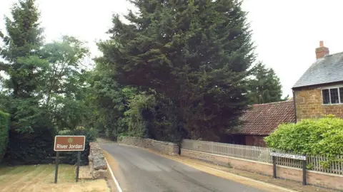 Rural bridge, showing road with stone wall where the bridge passes over the river and trees surrounding the road. A brown sign with white lettering to the left says "River Jordan" and a sign to the right indicates that we are Desborough Road. There is a stone-built house to the left.