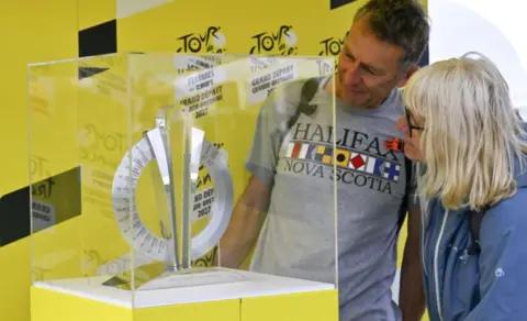 A woman with shoulder length blond hair and glasses, wearing a blue jacket and a man with short dark hair wearing a grey t-shirt with "Nova Scotia" written on it are both looking at the trophy which is on a yellow plinth and covered with a clear glass or plastic case. 