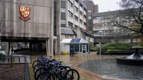 A grey, brutalist building on a rainy day. A red and gold Woking Borough Council logo is fixed to the side of the building and there are four bicycles locked to a railing outside the building.