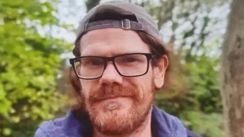 Shane Lynch is standing next to a tree and is smiling. He has ginger hair and a ginger beard and is wearing glasses and a back-to-front cap.