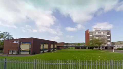 West Lakes Academy in Egremont. The school is behind a metal fence and is made of red brick. Two building's stand to the left while a large, five-storey building stands to the right.