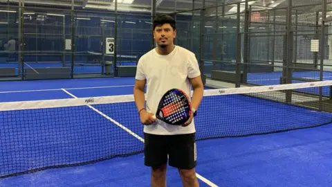 Umar is standing in front of a net on a blue-floored padel court. There are the glass walls and black railings of the court behind him. He is holding a black, red and blue padel racket in both hand. He is wearing a white T-shirt and black shorts that fall just above the knee. Umar has short black hair with a matching goatee and is smiling into the camera.