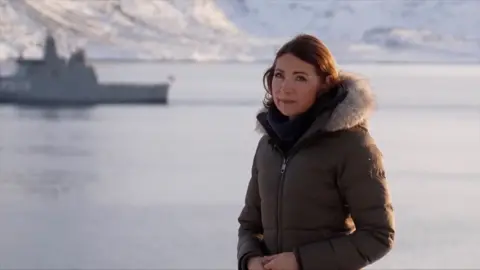 Katya Adler in a coat by the sea with a boat in the background.