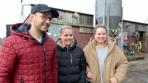 A man and two women wearing coats standing in front of a farm outdoors with cloudy skies behind.