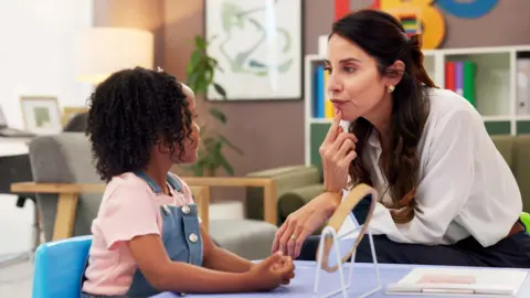 Getty Images The picture shows a female speech therapist with long brown hair pointing her index finger to her lips to demonstrate mouth movements.while sat a low table opposite a young girl. 